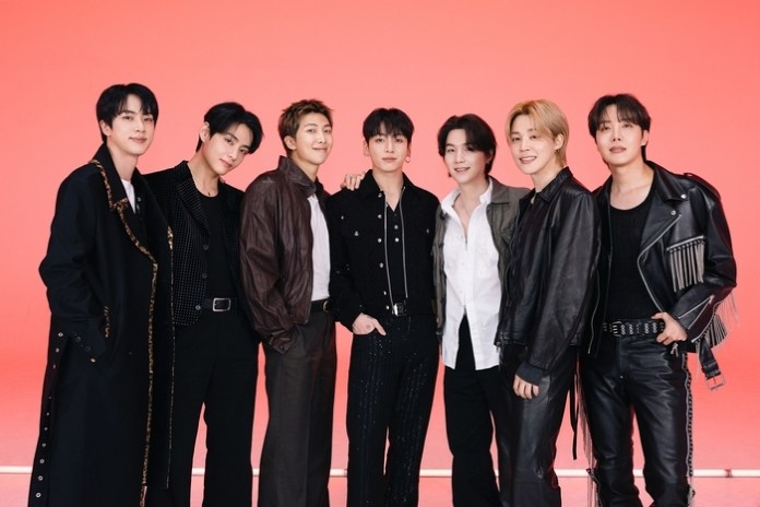 BTS standing together in front of a bright red background, each wearing stylish outfits.
