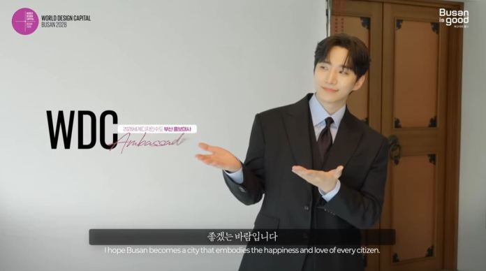 Lee Junho in a suit gestures with an inviting expression, promoting Busan as the World Design Capital 2028.