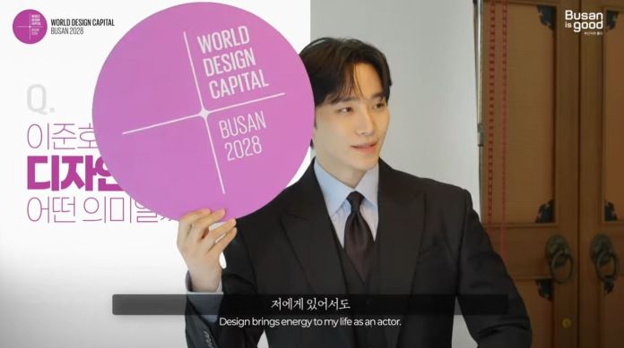 Lee Junho  in a formal suit holding a circular sign that reads 'WORLD DESIGN CAPITAL BUSAN 2028', with text in the background discussing the significance of design.