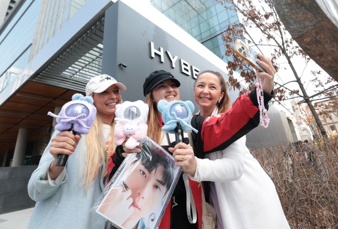 Three women smiling and taking a selfie outside a modern building with the word 'HYBE' visible, holding colorful lightsticks and a poster of a person.