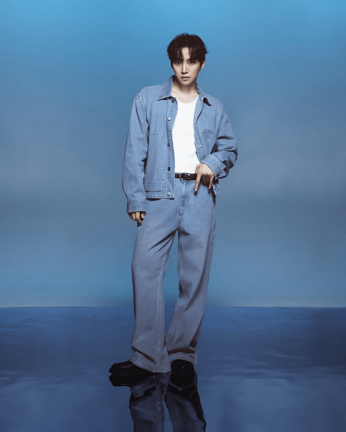 Lee Junho in a light denim outfit stands against a blue background. They are wearing a denim jacket and pants, paired with a white shirt, and are posing with one hand pointing down.