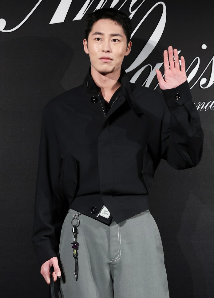 A man wearing a black jacket and gray pants, standing against a black backdrop, waving with a neutral expression.