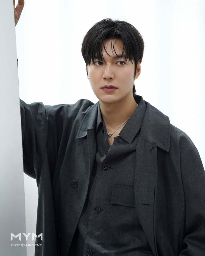 Lee Minho with dark hair and a serious expression, wearing a gray oversized jacket and shirt, poses against a white background.