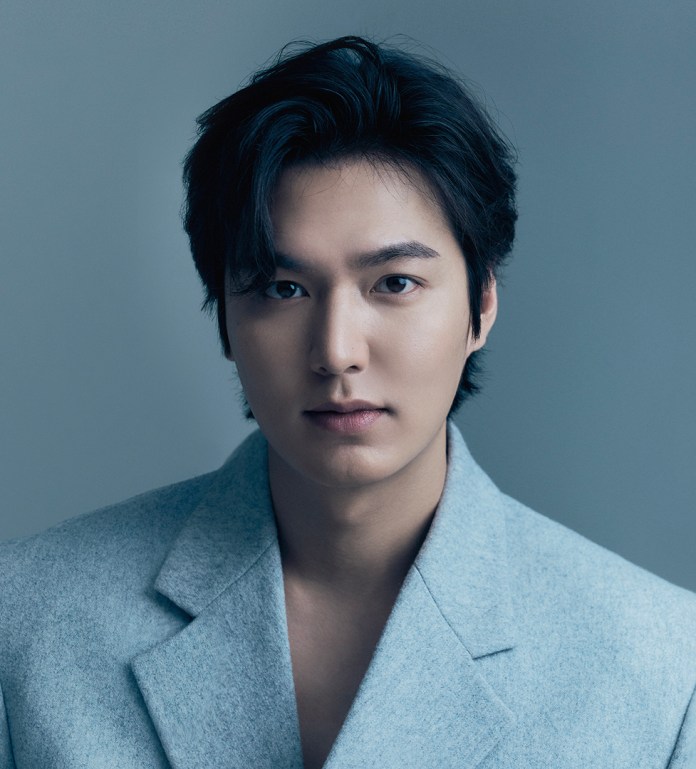 Lee Minho with dark hair, wearing a light gray oversized blazer, poses against a soft gray background, showcasing a serious expression.