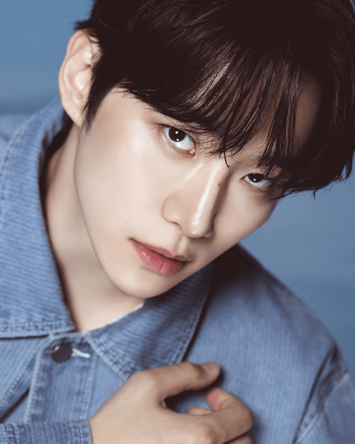 Lee Junho with short black hair, light skin, and a subtle expression. They are wearing a denim jacket and holding their hand to their collar, with a soft blue background.