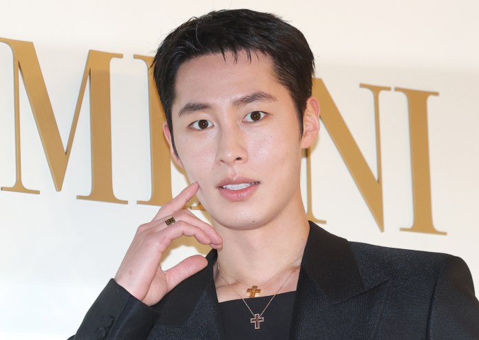 A young man with short black hair, wearing a black blazer and layered gold necklaces, poses with one hand near his face against a light-colored background featuring the word 'MARNI' in bold letters.