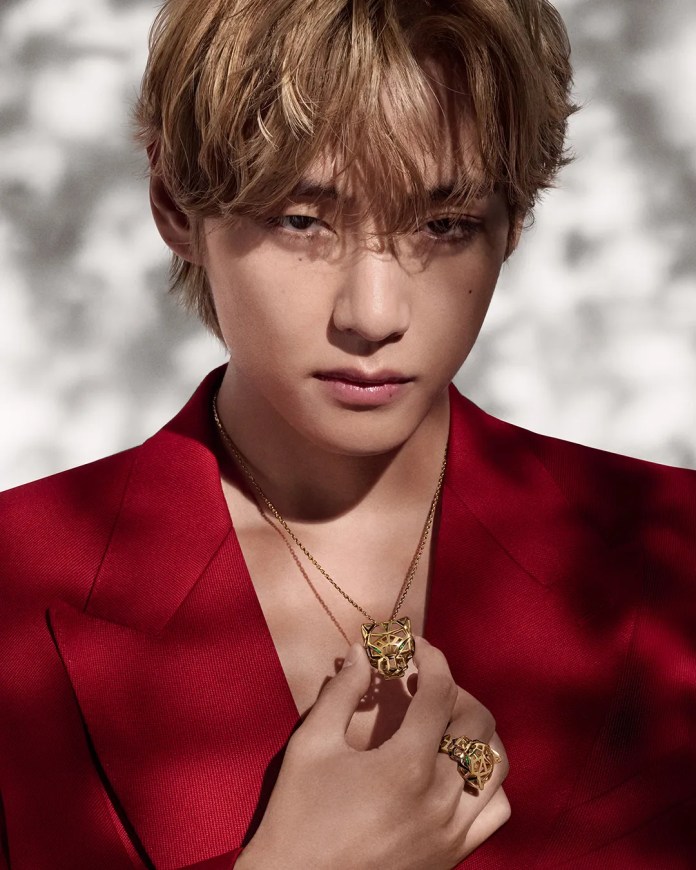 A close-up portrait of a young man with blonde hair, wearing a red suit. He holds a gold pendant and ring featuring a lion design, with a serious expression against a softly blurred background.