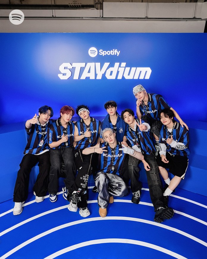 Stray Kids  members posing together in blue and black sports jerseys in front of a blue backdrop with the Spotify logo and the word 'STAYdium'.