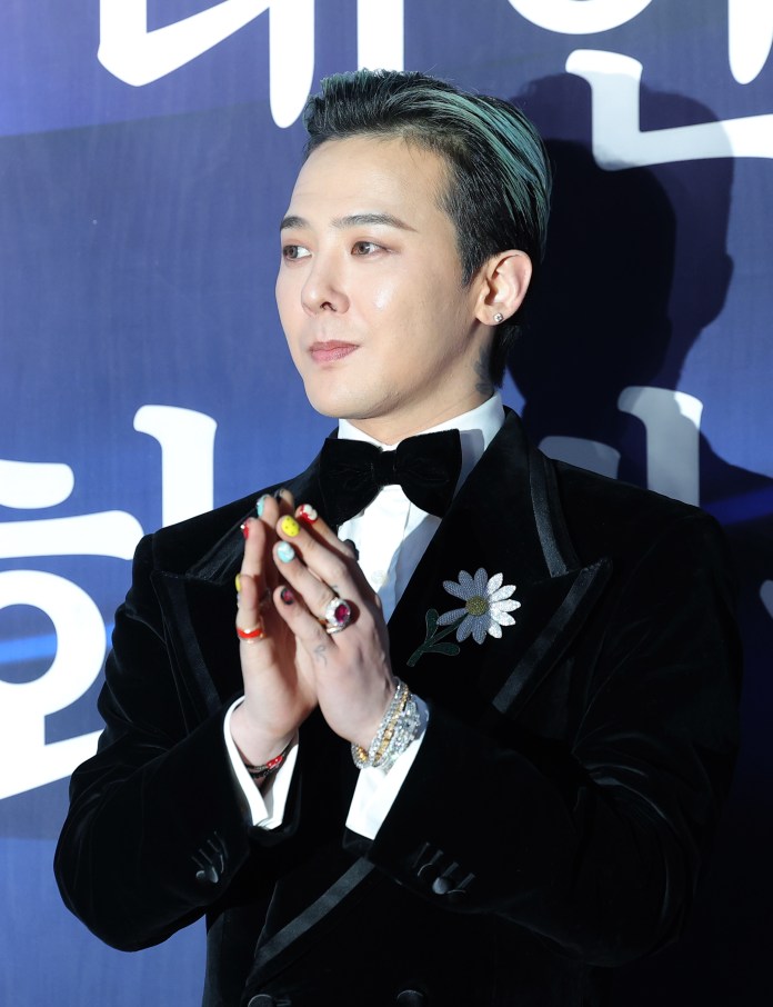 G-Dragon in a black velvet tuxedo with a white shirt and bow tie is smiling and clasping his hands together, adorned with colorful rings and nail polish. He has styled hair with light blue highlights and a flower brooch on his tuxedo. The background features Korean text.