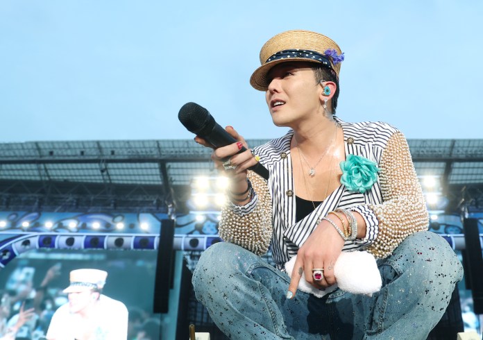 G-Dragon holding a microphone while sitting on stage, wearing a straw hat, a beaded jacket, and denim pants. Bright stage lights are visible in the background.