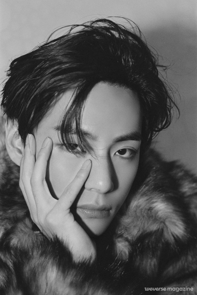 V with dark hair styled back, wearing a fur coat, gazing intently with one hand positioned near the face.