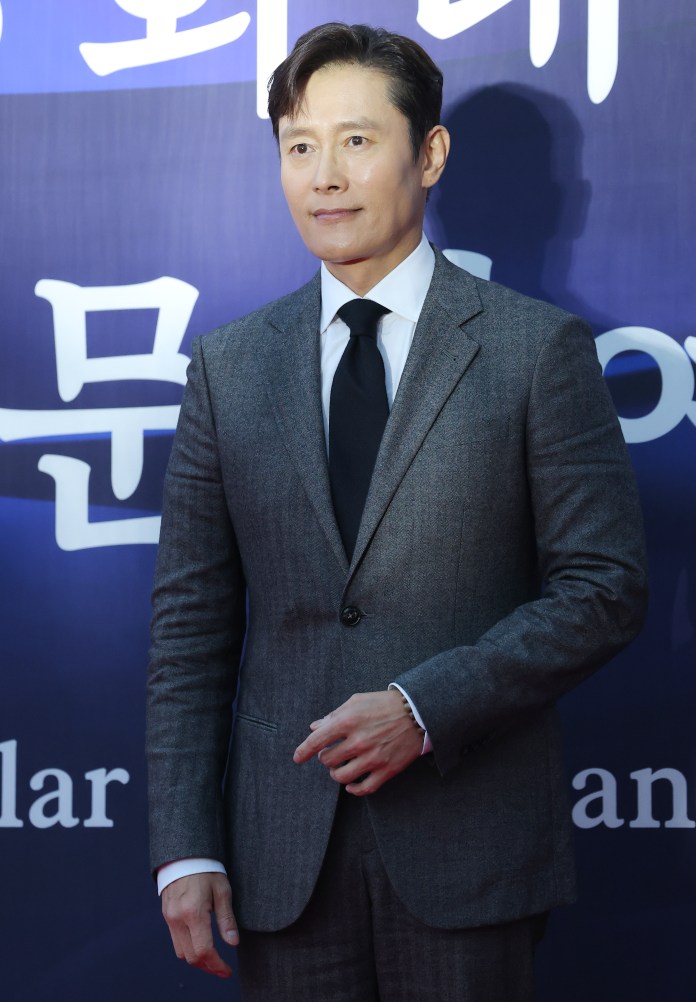 Lee Byung-hun in a grey suit standing in front of a blue and white backdrop with Korean text.