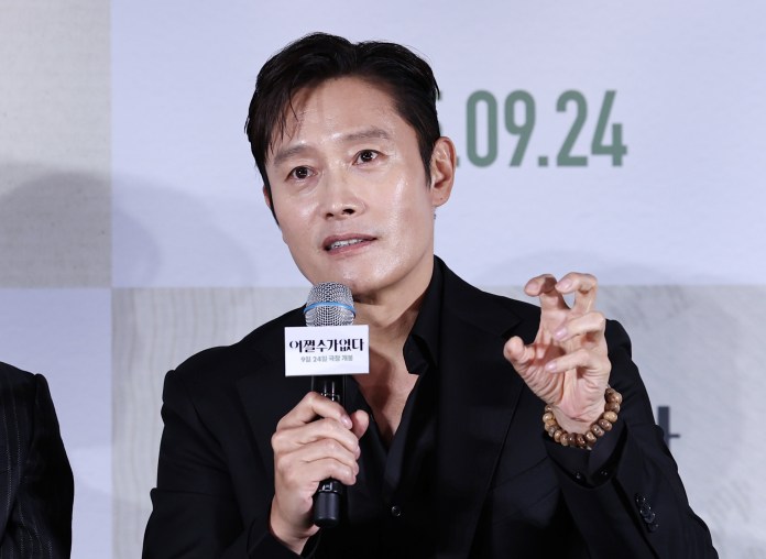 Lee Byung-hun in a black suit is speaking at an event, holding a microphone and gesturing with his hand.