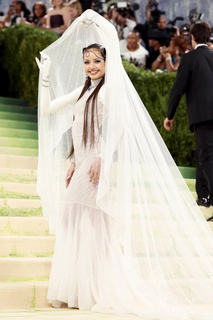 Lisa in a sparkling white dress and a long, sheer veil, smiling and waving at an event, surrounded by photographers and spectators.