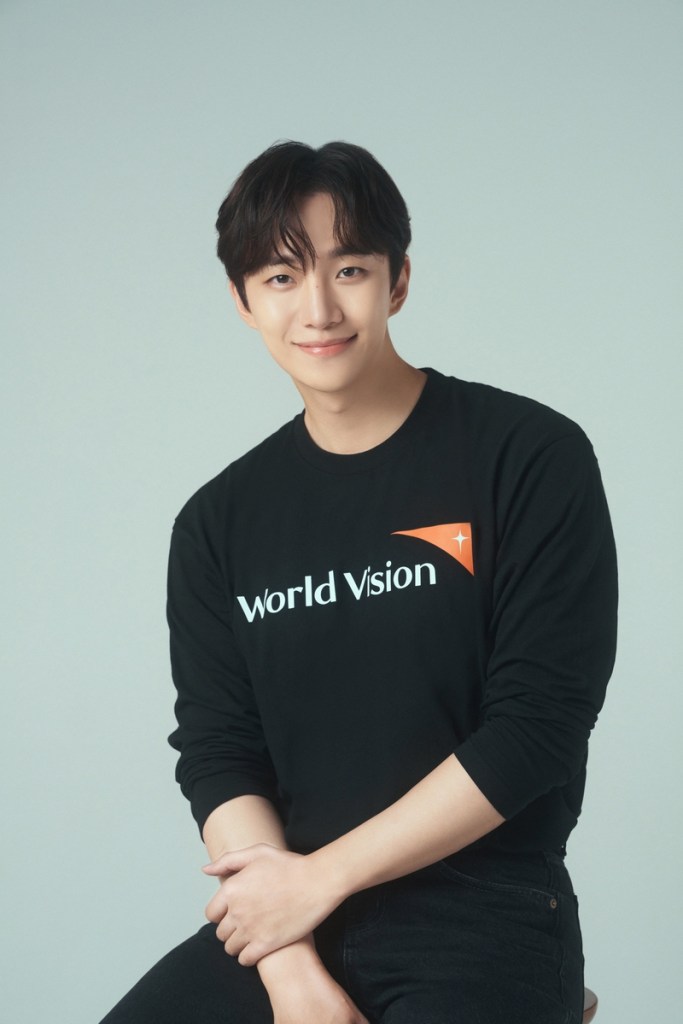 Lee Junho smiling while wearing a black long-sleeve shirt with the logo 'World Vision', sitting against a light gray background.