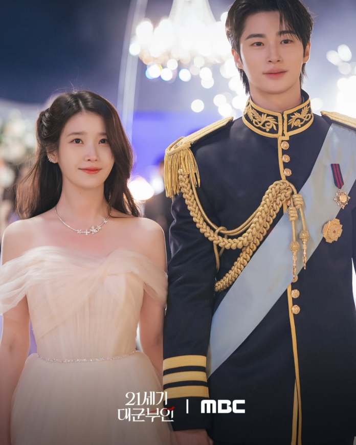 Byeon Woo-seok in a royal military uniform with gold embellishments stands beside IU in a flowing off-the-shoulder gown, both smiling at an event with shimmering lights in the background.