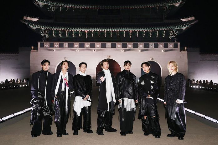 BTS dressed in stylish, modern black outfits stands in front of a traditional architectural structure at night, with soft lighting illuminating the scene.