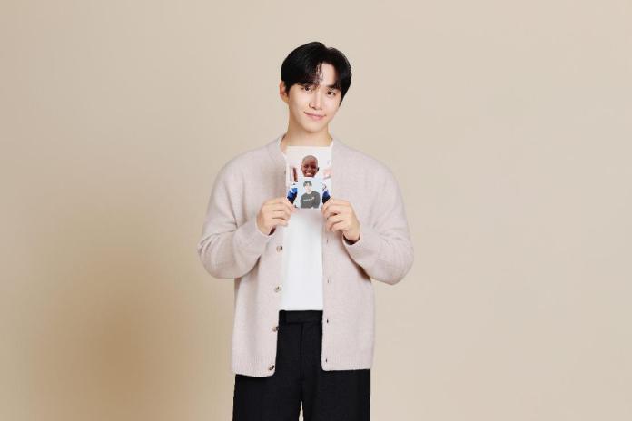 Lee Junho wearing a light cardigan and holding a photo is smiling at the camera against a neutral background.