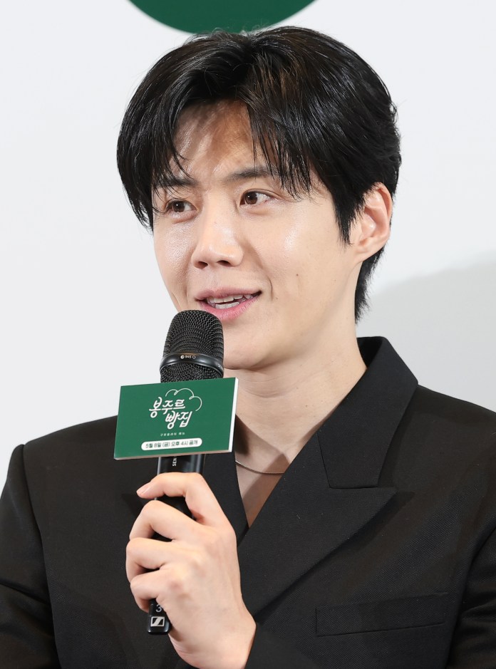 Kim Seon-ho with dark hair speaking into a microphone at an event, wearing a black blazer.