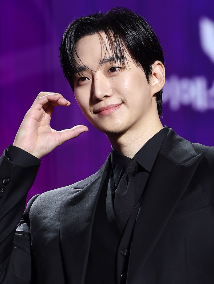 Lee Junho in a black suit smiling and making a heart shape with their hand against a colorful background.