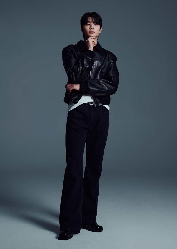 Kim Jaewon with one hand on his chin, wearing a black leather jacket, white t-shirt, and dark trousers against a gray background.