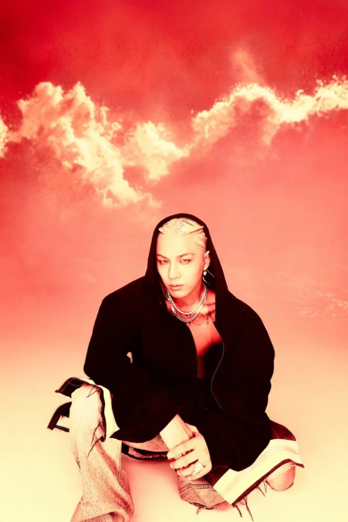 Taeyang sitting on the ground, dressed in a hooded black jacket and distressed jeans, against a vibrant red background with swirling clouds.