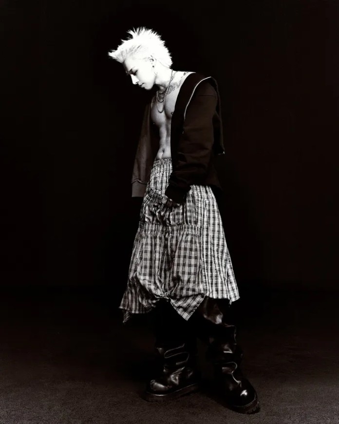 Taeyang with spiky white hair wearing a cropped black hoodie and a plaid skirt, standing in profile against a dark background, exuding an edgy fashion vibe.