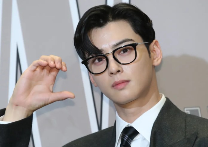 Cha Eun-woo in a suit and glasses poses, making a hand gesture as if illustrating a small size.