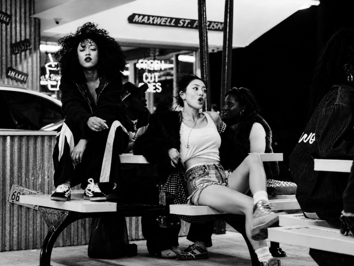 Jenevieve and Jihyo sitting on a bench at night, dressed in trendy urban clothing, with vibrant neon signs in the background.