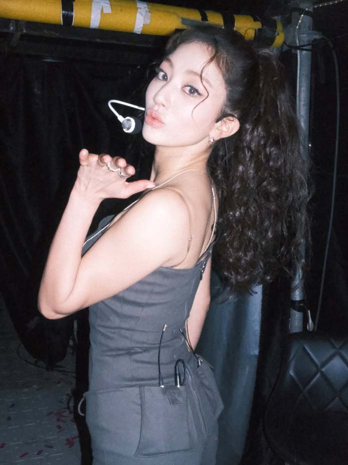 Jihyo with curly hair wearing a fitted gray outfit, facing away from the camera, blowing a kiss. She has a microphone attached and stands against a dark background.