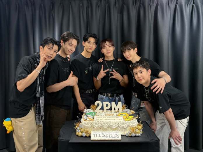 2PM members posing together in front of a decorated cake celebrating the 18th anniversary of a concert. They are all wearing black shirts and smiling, with one member making a peace sign.