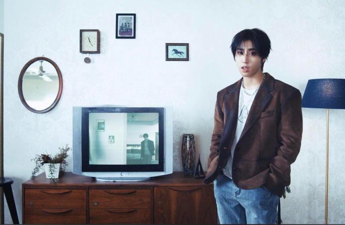 Han standing in a retro-styled living room, wearing a brown blazer and casual attire, with a vintage television displaying a reflection of another person. The room features patterned wallpaper, a mirror, and various framed pictures on the wall.
