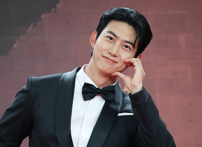 Taecyeon in a tuxedo making a playful gesture with his hand while smiling at the camera.