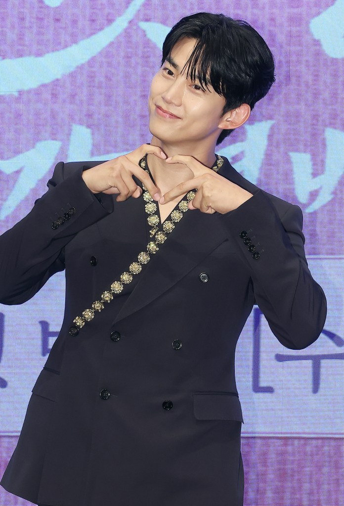 Taecyeon in a dark double-breasted suit making a heart shape with his hands while smiling for the camera.