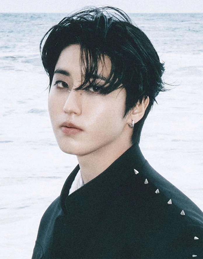 Han with tousled black hair and a piercing, wearing a black jacket adorned with small metallic studs, gazes directly at the camera with a serene ocean background.