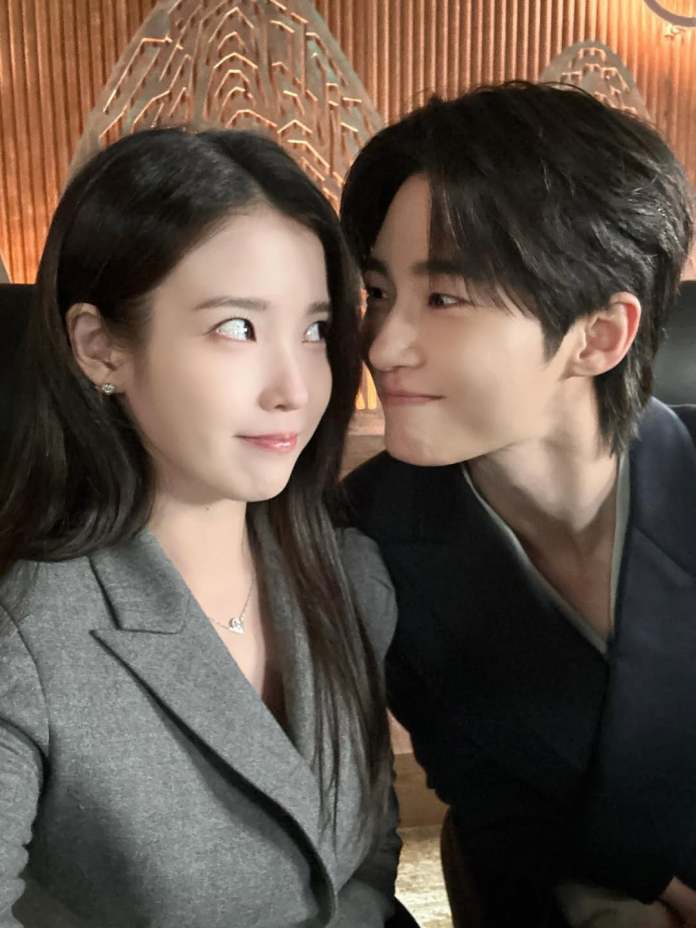 A close-up selfie of IU and Byeon Woo-seok, IU with long dark hair wearing a gray blazer and Byeon Woo-seok with short dark hair wearing a dark outfit, sharing a playful gaze at each other.