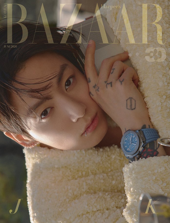 Jungkook with dark hair, resting his head on his hand while wearing a fuzzy yellow sweater and a blue watch. The image features intricate tattoos on his hand, and he has a piercing in his lower lip.