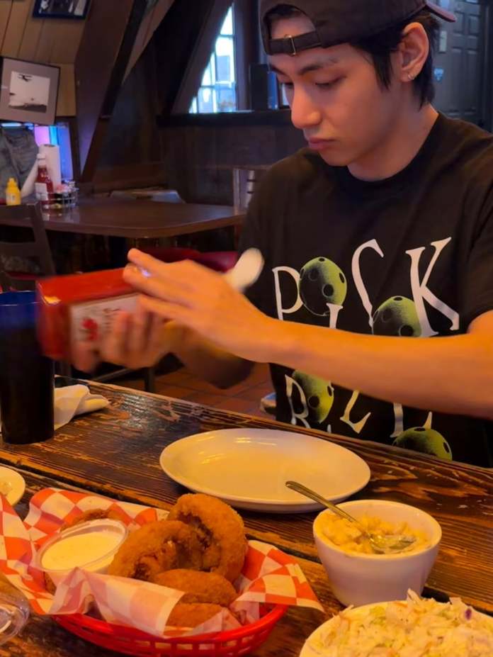 V sitting at a table in a restaurant, preparing to add ketchup to a plate. A basket of onion rings, a bowl of macaroni and cheese, and a side of coleslaw are on the table.