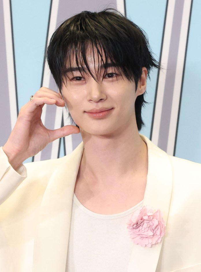 Byeon Woo-seok with dark, wet hair smiling and making a hand gesture. He is wearing a white jacket and a white shirt, accessorized with a pink flower pin.