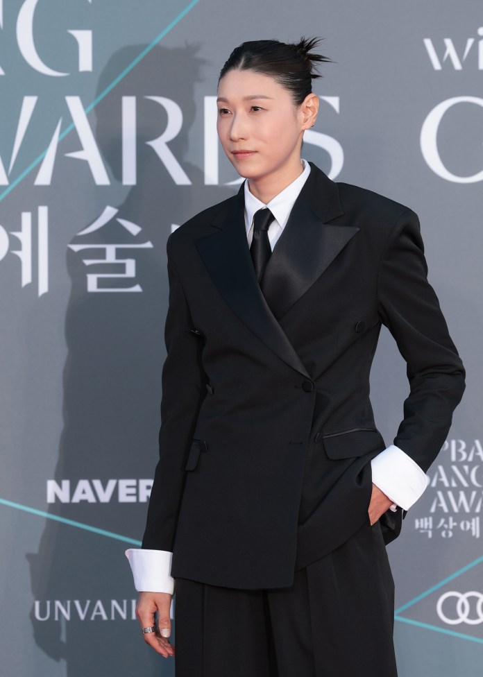 Kim Yeon-koung wearing a stylish black suit with a white shirt and tie, standing confidently at a formal event backdrop.