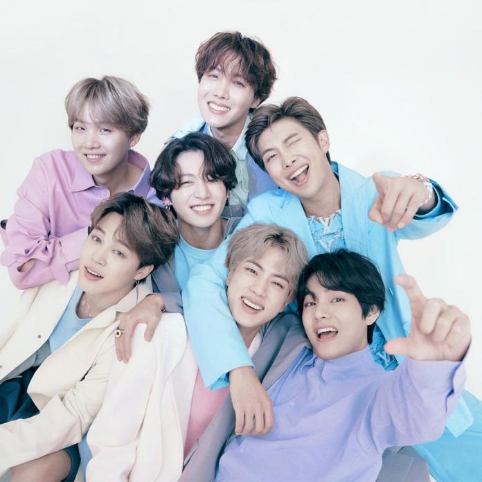 BTS smiling together, wearing pastel-colored outfits, posing happily against a light background.