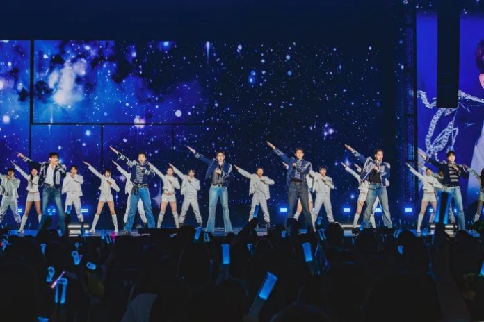 2PM on stage during concert, posing dynamically against a backdrop of a starry sky and colorful lights, with fans holding light sticks in the foreground.