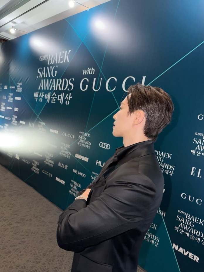 Side profile of Lee Junho in a black suit at the 62nd Baek Sang Awards, with a backdrop featuring the event name and sponsors including Gucci.