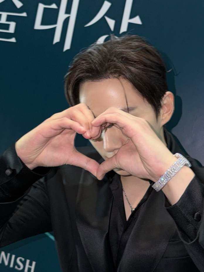 Lee Junho wearing a black shirt makes a heart shape with their hands, showcasing a sparkling bracelet.
