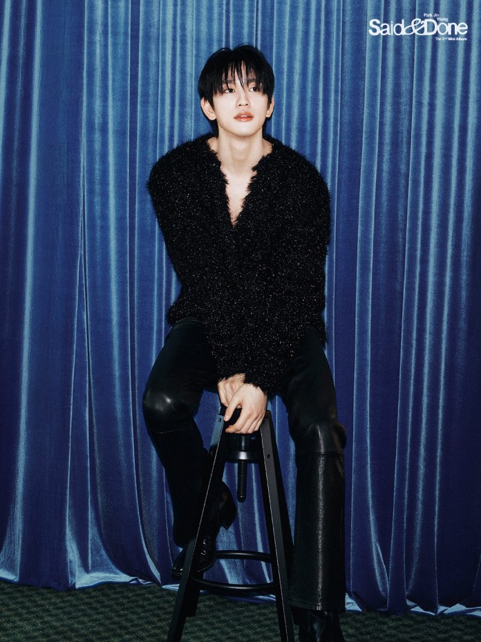 Park Jinyoung  sitting on a stool against a blue curtain backdrop, wearing a black fuzzy sweater and leather pants, with a thoughtful expression.