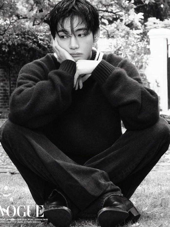 BTS V in a black sweater and dark jeans sits cross-legged on the grass, resting his chin on his hand with a thoughtful expression. The image is in black and white, featuring soft, natural surroundings.