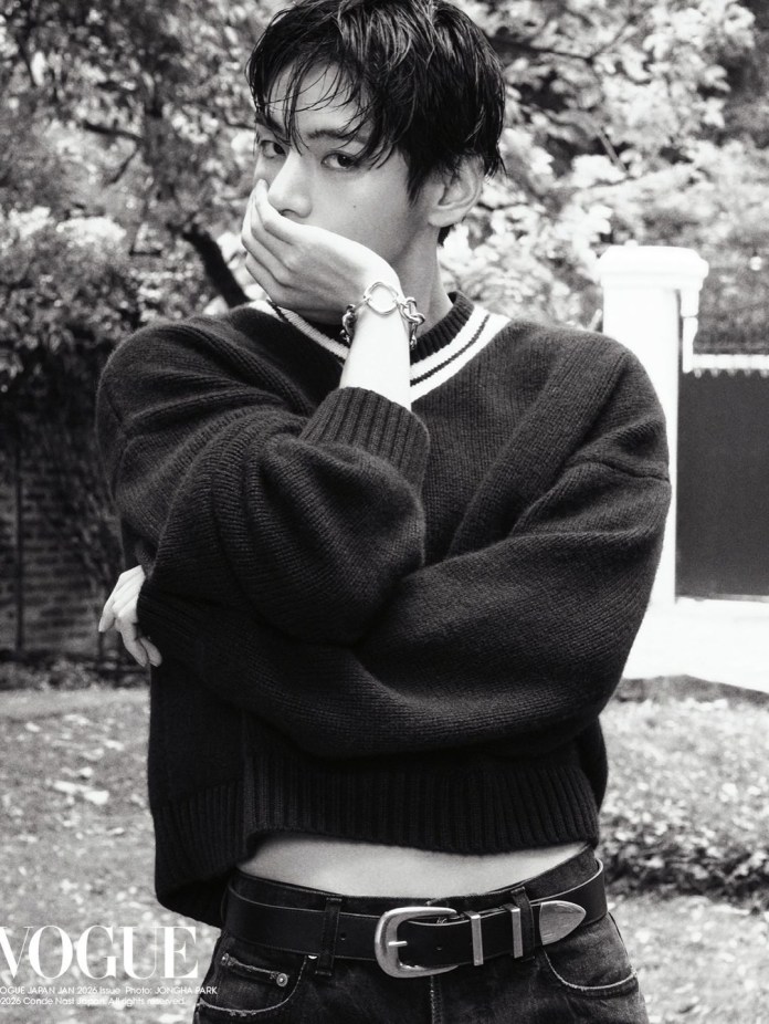 BTS V in a black and white image wearing a cropped black sweater with a white collar, holding their hand to their mouth in a thoughtful pose, standing outdoors with blurred greenery in the background.