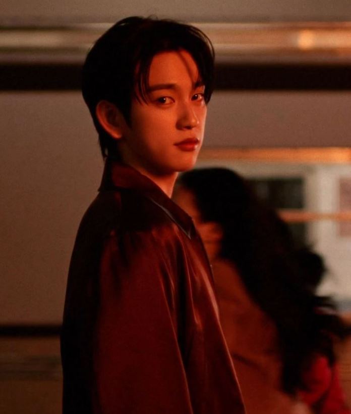 Park Jinyoung with dark hair wearing a shiny red shirt, looking over their shoulder against a softly lit background.