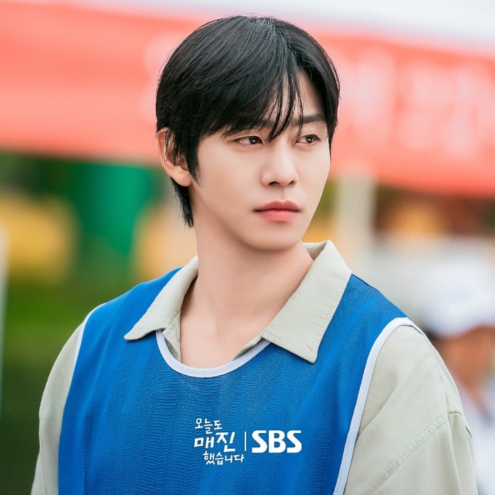Ahn Hyo-seop with dark, medium-length hair is wearing a blue vest over a light-colored shirt, looking thoughtfully into the distance. The background features blurred greenery and colorful banners.