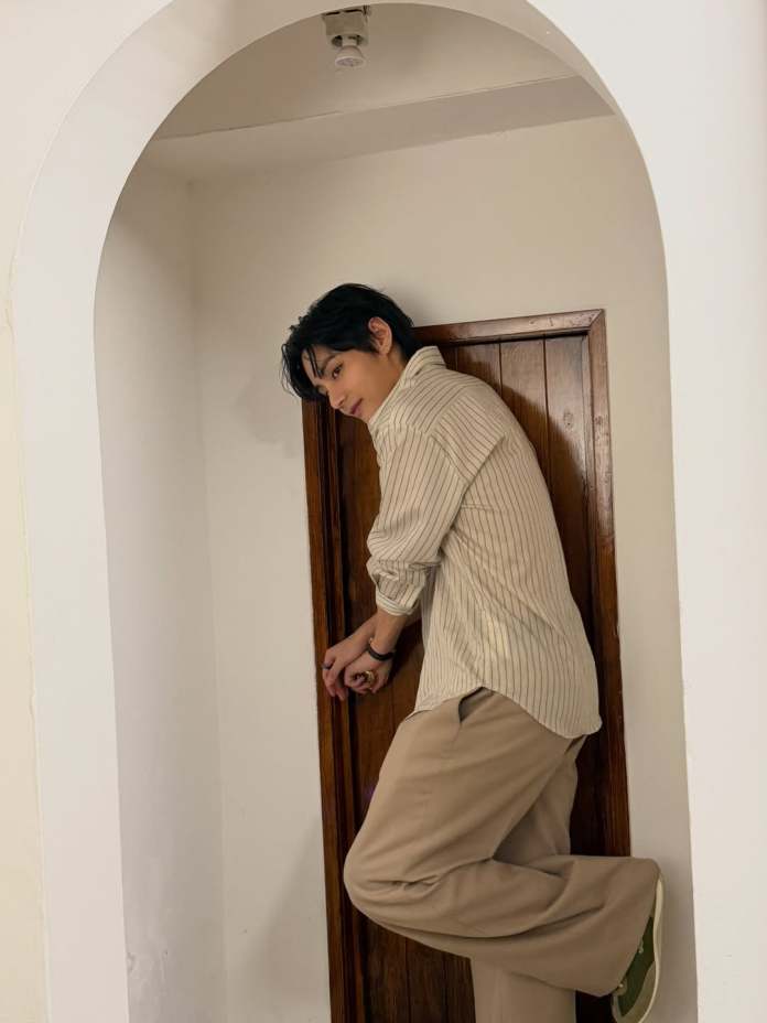 A person leaning against a wooden door within an arched doorway, wearing a striped shirt and beige pants.